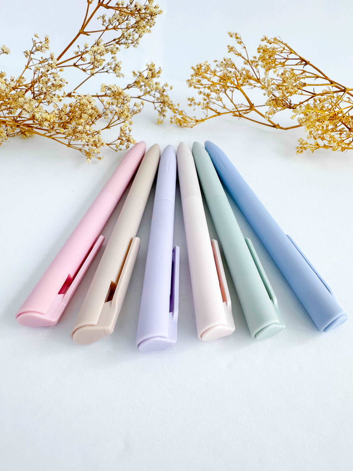 Six pastel-colored slim pens or stylus pens arranged in a fan shape on a white surface, with dried floral branches in the background. The pens are pink, beige, lavender, blush, mint green, and light blue.