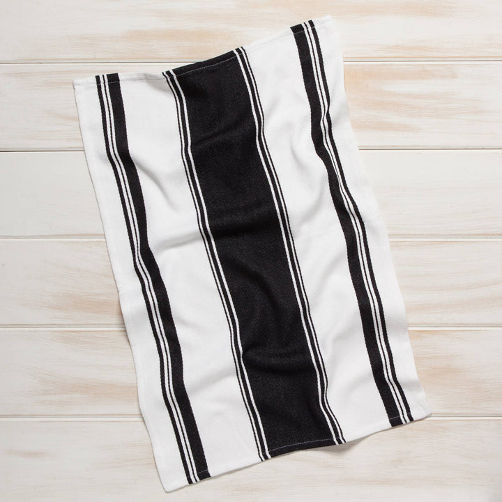 A white cotton kitchen towel with bold black vertical stripes, laid flat on a light-colored wooden plank surface.