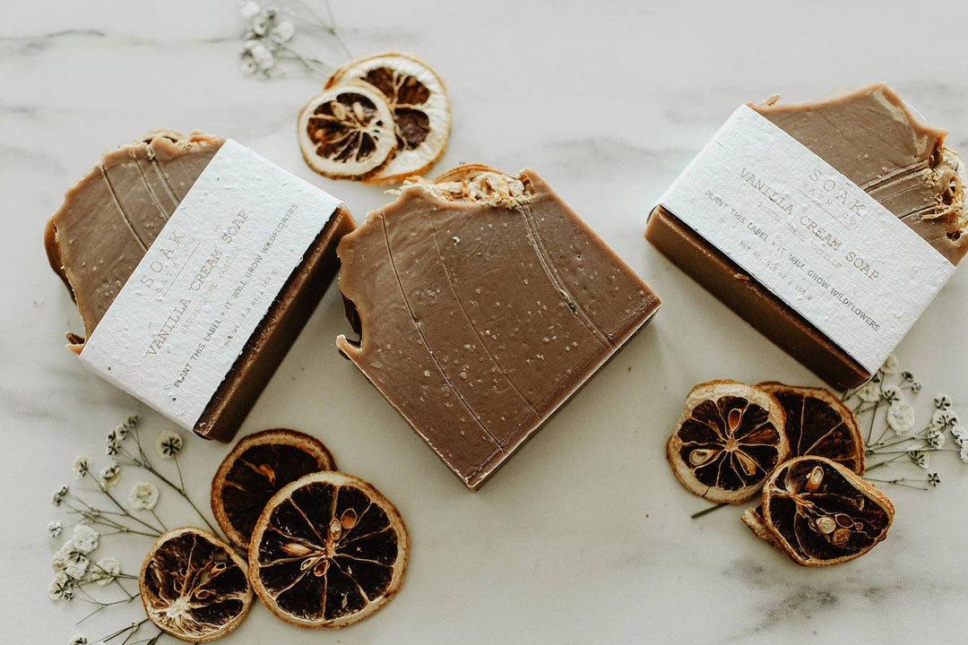 Three bars of soap with white labels on a marble surface with dried oranges and flowers.