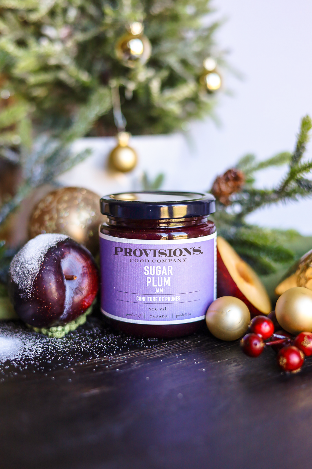 A jar of Provisions Farm Company Sugar Plum condiment and preserves sits on a dark tabletop surrounded by holiday decorations, including a sugar-dusted plum, gold ornaments, and evergreen branches.