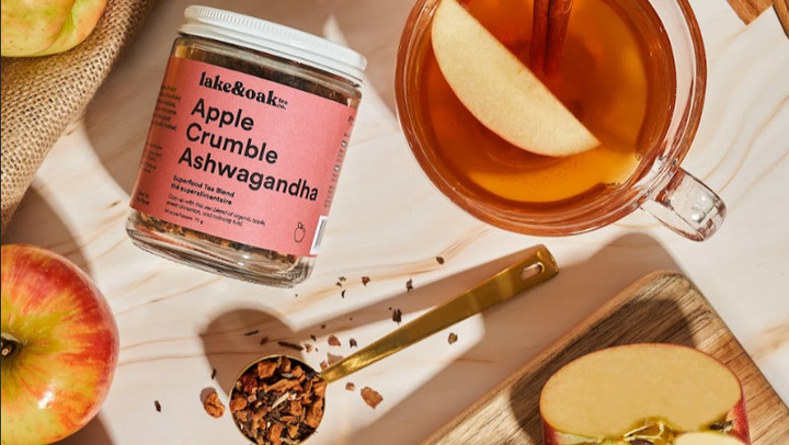 A jar of Lake &amp; Oak Apple Crumble Ashwagandha tea placed on a light surface with whole and sliced apples, a glass mug of amber tea with an apple slice, and a spoonful of loose tea grounds.