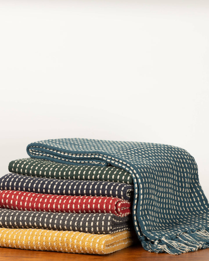A stack of folded woven throw blankets in teal, green, navy, red, charcoal, and mustard yellow, each with a white dotted pattern, displayed on a wooden surface against a plain white background.