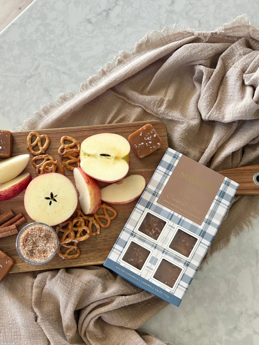 A boxed assortment of chocolate squares and caramel treats sits on a wooden board with sliced red apples, pretzels, cinnamon, and a beige linen cloth on a light stone surface.