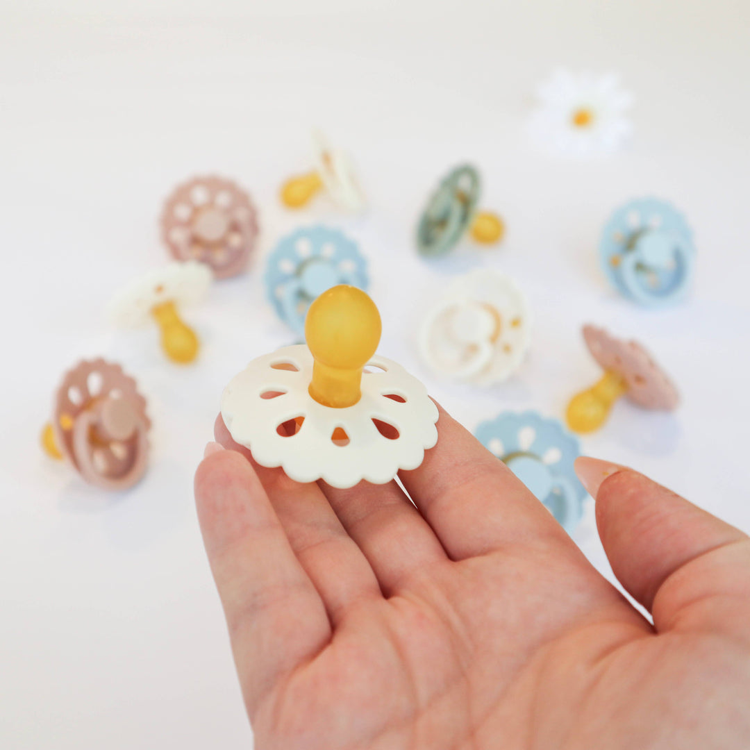 Hand holding a white pacifier with colorful pacifiers in the background on a light surface.