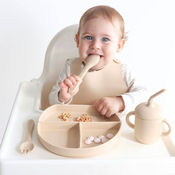 Baby in a high chair with wooden utensils and a plate of food on a white background