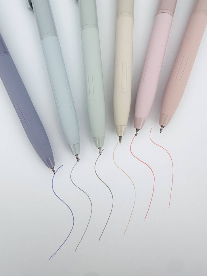 Six pastel-colored retractable pens in lavender, light blue, mint, cream, blush pink, and dusty rose, arranged diagonally on a white background with their ink lines fanning out below.