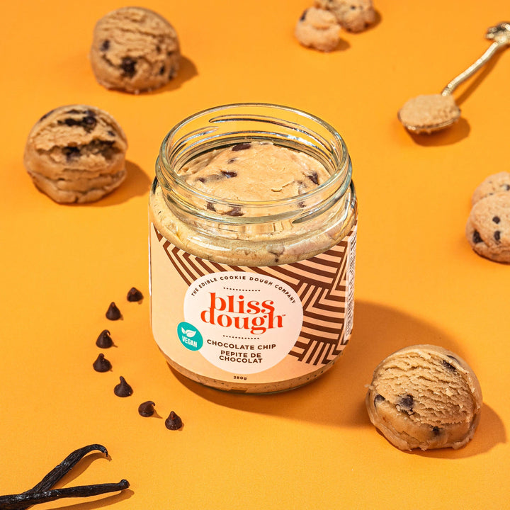 Jar of 'bliss dough' chocolate chip cookie dough on an orange background with cookies and vanilla beans.