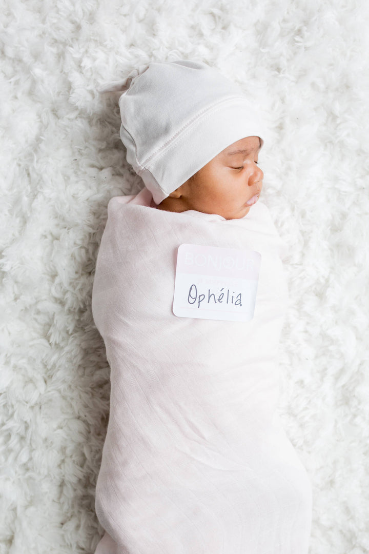 Newborn baby swaddled in a light pink blanket with a name tag on a white fluffy surface