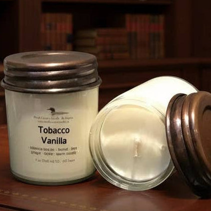 Candle, bottle of Cremmed, and smoking cigar on a wooden surface with a bookshelf in the background.