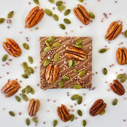 A square chocolate bar decorated with drizzle and diagonal stripes, topped with pecans and green pumpkin seeds, arranged on a white background with scattered pecans and seeds.