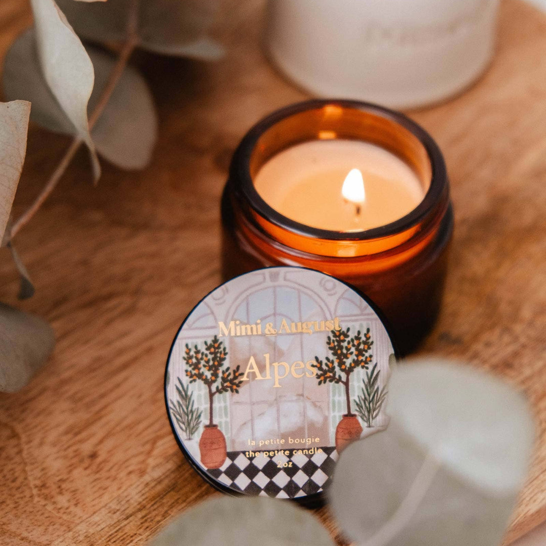 Candle in an amber jar with a decorative label on a wooden surface surrounded by greenery