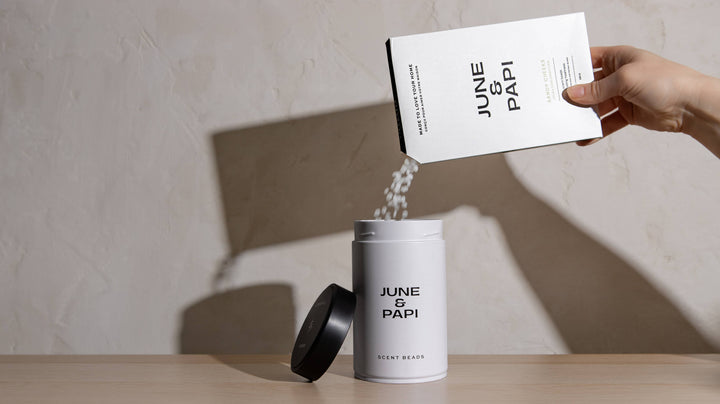A hand pours white granules from a white box labeled JUNE &amp; PAPI into an open white cylindrical tin with a black lid, set on a light wood surface against a textured beige wall.