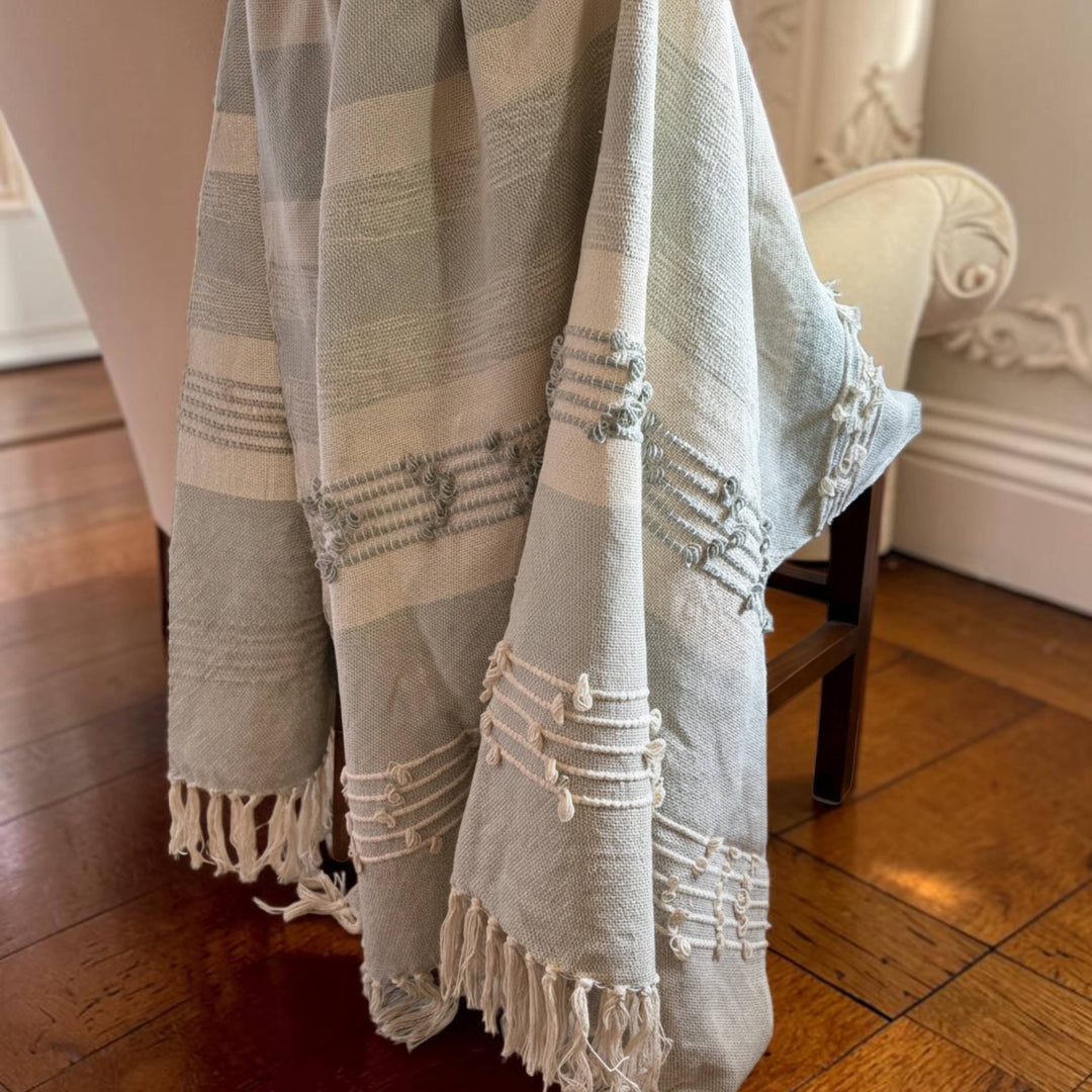 A woven throw blanket with horizontal stripes in light blue-gray and cream, draped over the back of a chair in a room with hardwood floors and white walls.