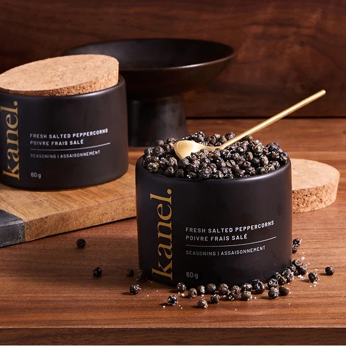 Two black glass jars of Kanel fresh salted peppercorns with cork lids on a wooden table, with one jar open to show the peppercorns and a small gold spoon resting inside.