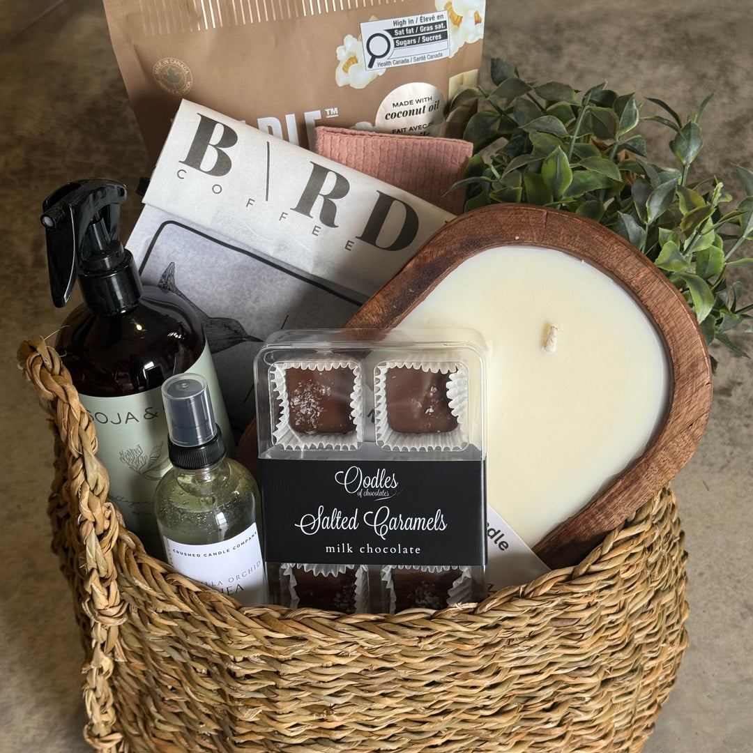 A woven straw gift basket filled with assorted gift items, including a large cream-colored candle in a wooden bowl, a package of salted caramels, a brown coffee bag, a spray bottle, and a small green bottle, arranged on a concrete floor.